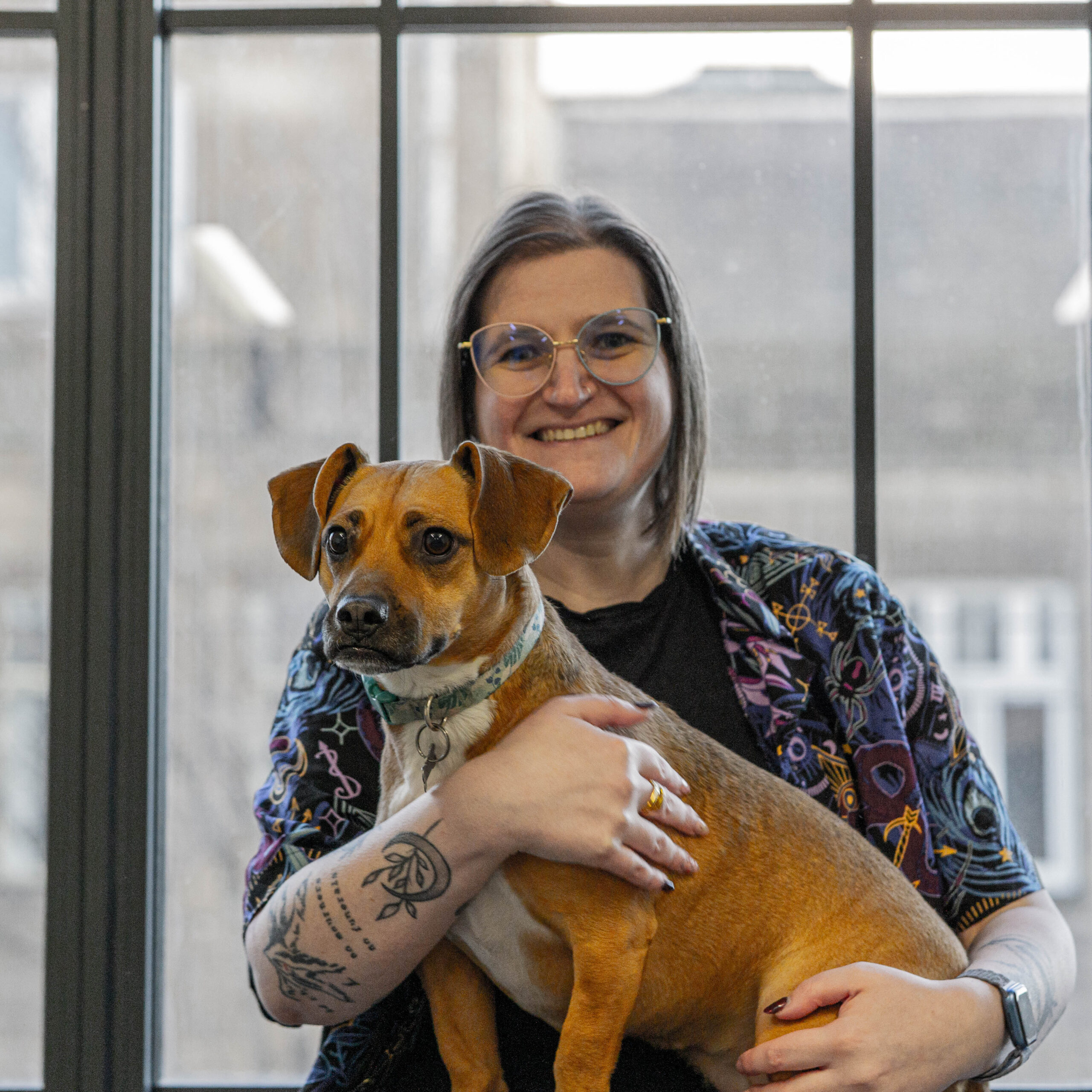 KGT sits with their dog, Jester, on their lap in front of the bright windows of the office